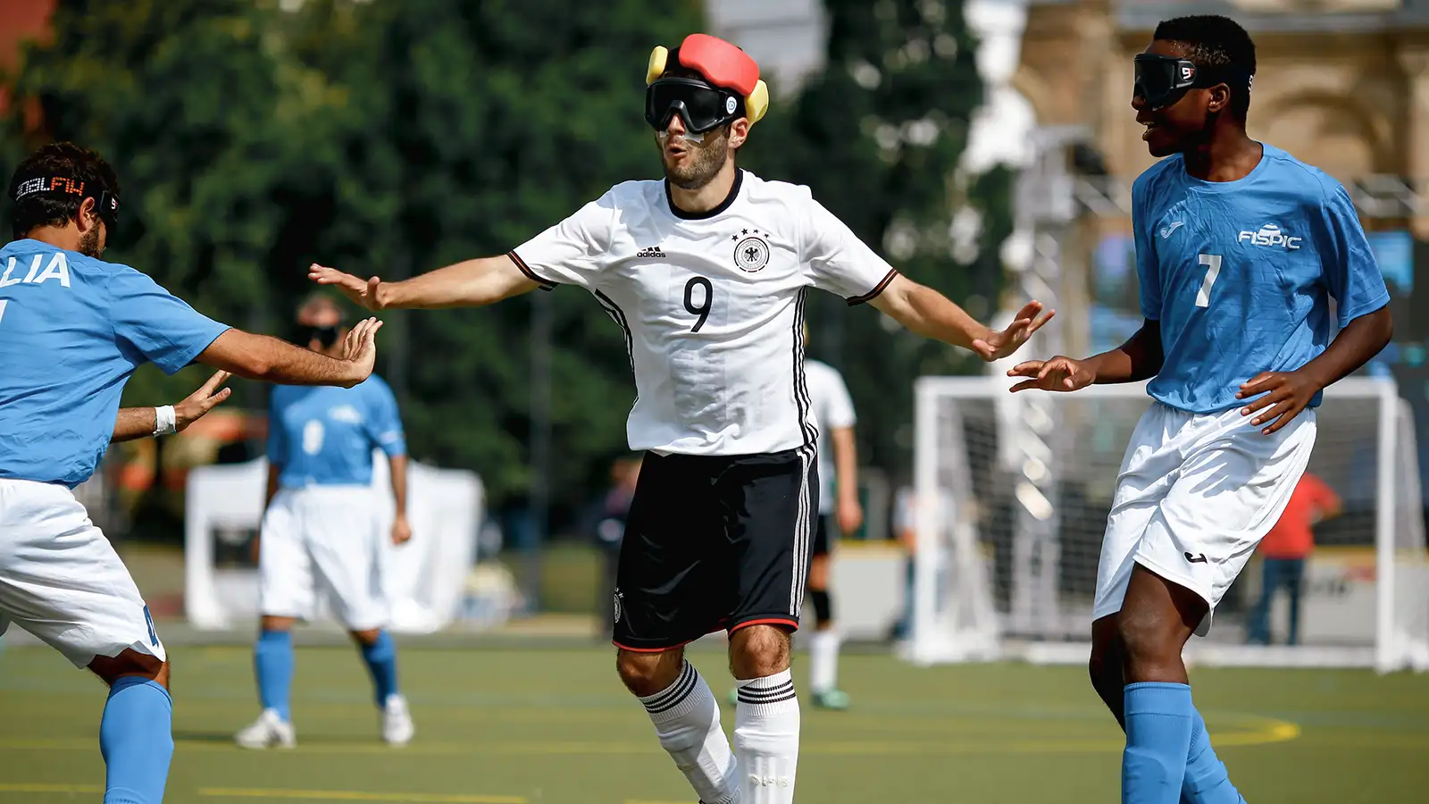 3 Blindenfußballspieler, 2 in blauen Trikots und einer in weiß, auf dem Spielfeld. Der Spieler in weiß kontrolliert den Ball, alle tragen Augenbinden.