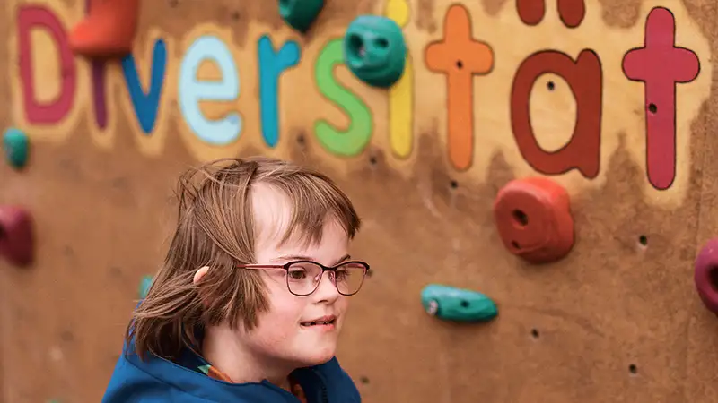 Ein Kind mit Down-Syndrom lächelt vor einer Kletterwand mit dem Schriftzug "Diversität" im Hintergrund.