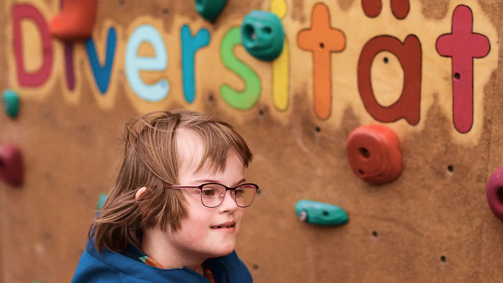 Ein Kind mit Down-Syndrom lächelt vor einer Kletterwand mit dem Schriftzug "Diversität" im Hintergrund.