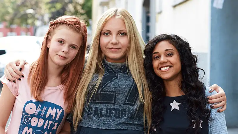 Drei Mädchen unterschiedlichen ethnischen Hintergrunds sitzen draußen und lächeln in die Kamera.