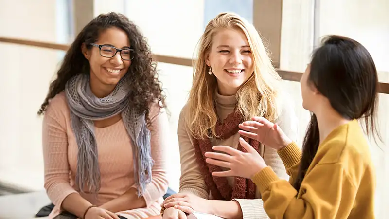Drei junge Frauen unterschiedlichen ethnischen Hintergrunds sitzen zusammen und unterhalten sich lachend in einem hellen Raum.