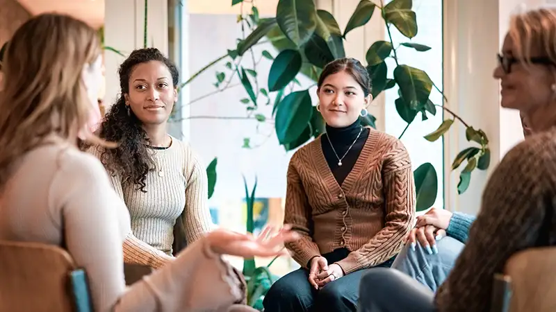 Eine Gruppe von vier Frauen unterschiedlichen ethnischen Hintergrunds sitzt in einem Kreis in einem Raum mit Pflanzen und unterhält sich.