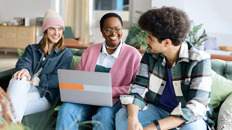 Drei junge Erwachsene unterschiedlichen ethnischen Hintergrunds auf einem Sofa in einem Wohnzimmer. Sie lachen, während sie auf einen Laptop schauen.