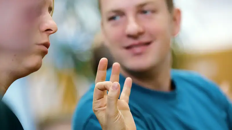 Zwei Personen kommunizieren in Gebärdensprache, wobei der Fokus auf den Händen liegt, die ein Zeichen formen.