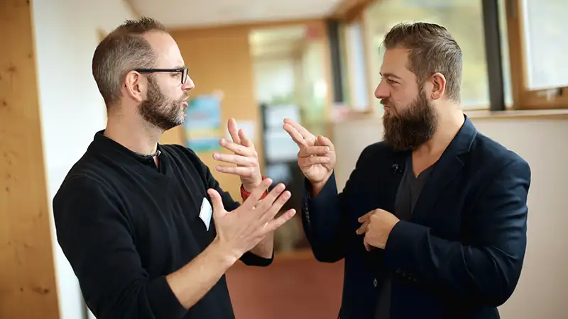 Zwei Männer unterhalten sich in Gebärdensprache in einem hellen Korridor, beide gestikulieren aktiv mit ihren Händen.