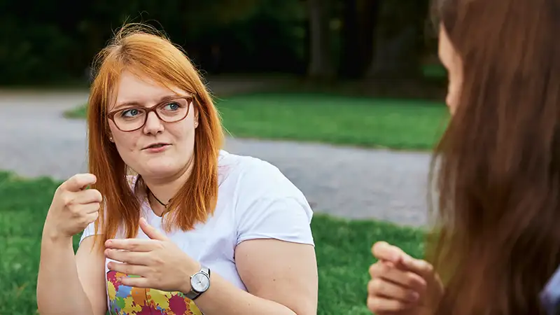 Zwei Frauen unterhalten sich draußen in Gebärdensprache, eine von ihnen hat rote Haare und trägt eine Brille.