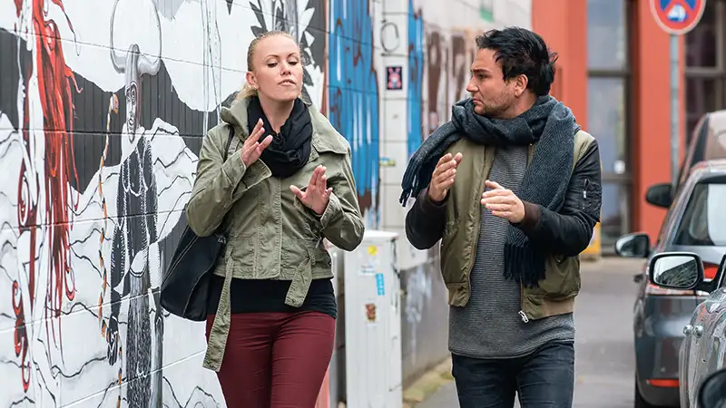 Zwei Personen unterhalten sich in Gebärdensprache, während sie auf einem Gehweg entlang einer mit Graffiti bedeckten Wand spazieren.