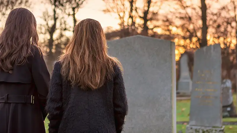 Zwei Frauen stehen bei Sonnenuntergang auf einem Friedhof und schauen auf einen Grabstein.