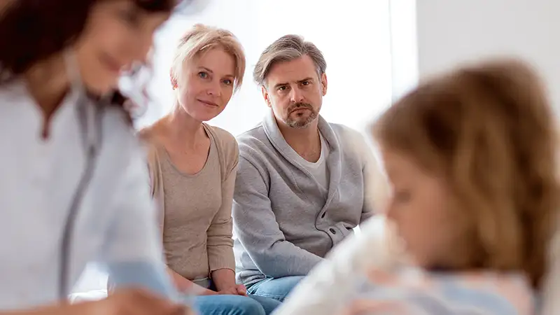 Ein Kind wird im Krankenhaus von einer Ärztin untersucht. Im Hintergrund sind die Eltern mit besorgtem Blick.