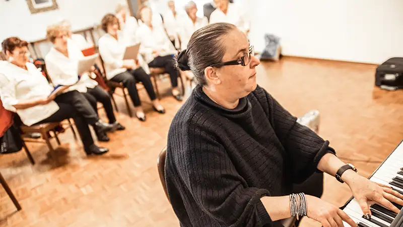 Eine Frau spielt Klavier, während eine Gruppe älterer Menschen im Hintergrund in einem Raum sitzt und dabei singt.