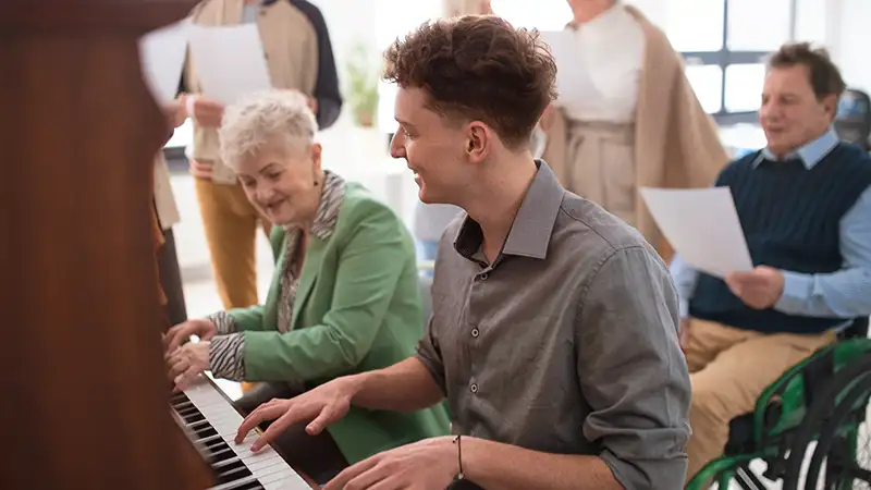 Ein junger Mann spielt Klavier und eine ältere Frau lächelt neben ihm. Im Hintergrund singen weitere ältere Menschen.