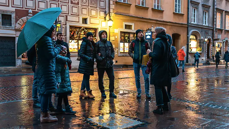Eine Gruppe von Menschen bei Regen auf einer beleuchteten Straße in einer historischen Stadt. Einige haben Regenschirme. Sie hören einem Führer zu.
