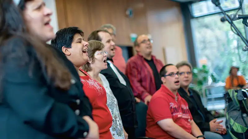 Eine Gruppe von Menschen unterschiedlichen Alters singt zusammen in einem Raum, einige sitzen, andere stehen.