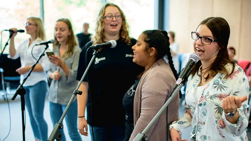 Eine Gruppe junger Frauen singt in Mikrofone in einem Raum, alle lächeln und wirken engagiert.