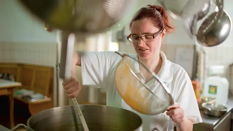 Eine Frau mit Brille und rotem Haar kocht in einer Großküche und rührt in einem großen Topf.