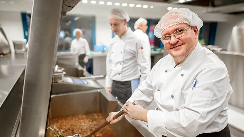 Ein lächelnder Koch in weißer Uniform rührt in einem großen Topf mit Soße in einer professionellen Küche, während ein Kollege im Hintergrund arbeitet.