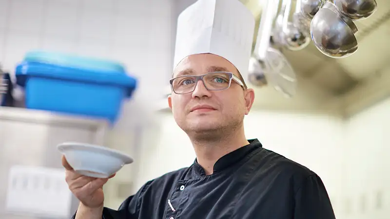 Ein Koch mit weißer Mütze hält Schüssel in einer Küche.
