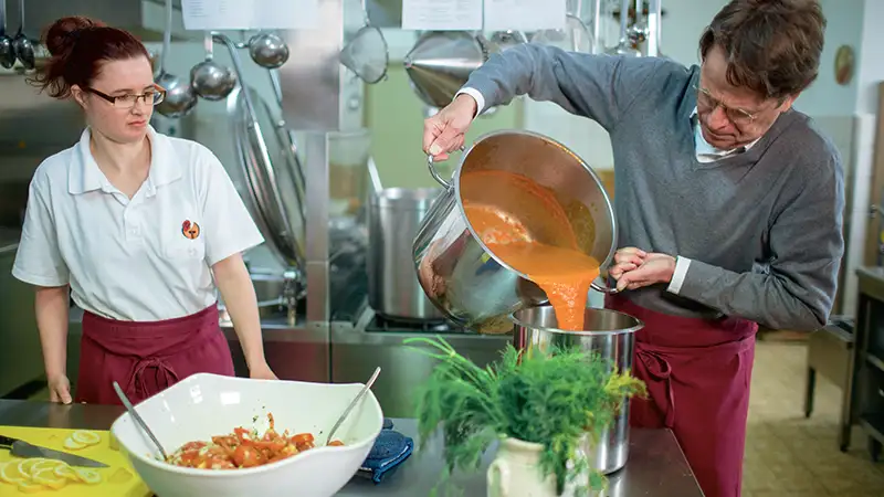 Situation in einer Großküche: Ein Mann gießt Sauce über Gemüse, während eine Frau zusieht
