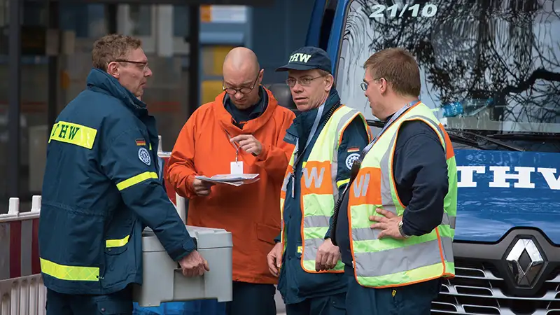 Vier Arbeiter in Warnwesten führen eine Besprechung vor einem LKW des Technischen Hilfswerks durch.