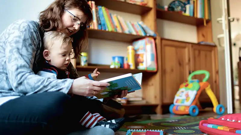 Eine Frau liest einem Kleinkind ein Buch in einem Spielzimmer vor.