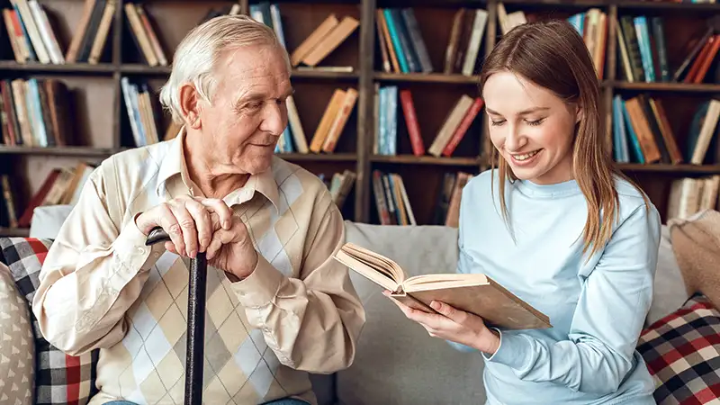 Eine junge Erwachsene liest einem älteren Mann ein Buch vor. Im Hintergrund ist ein großes Bücherregal.