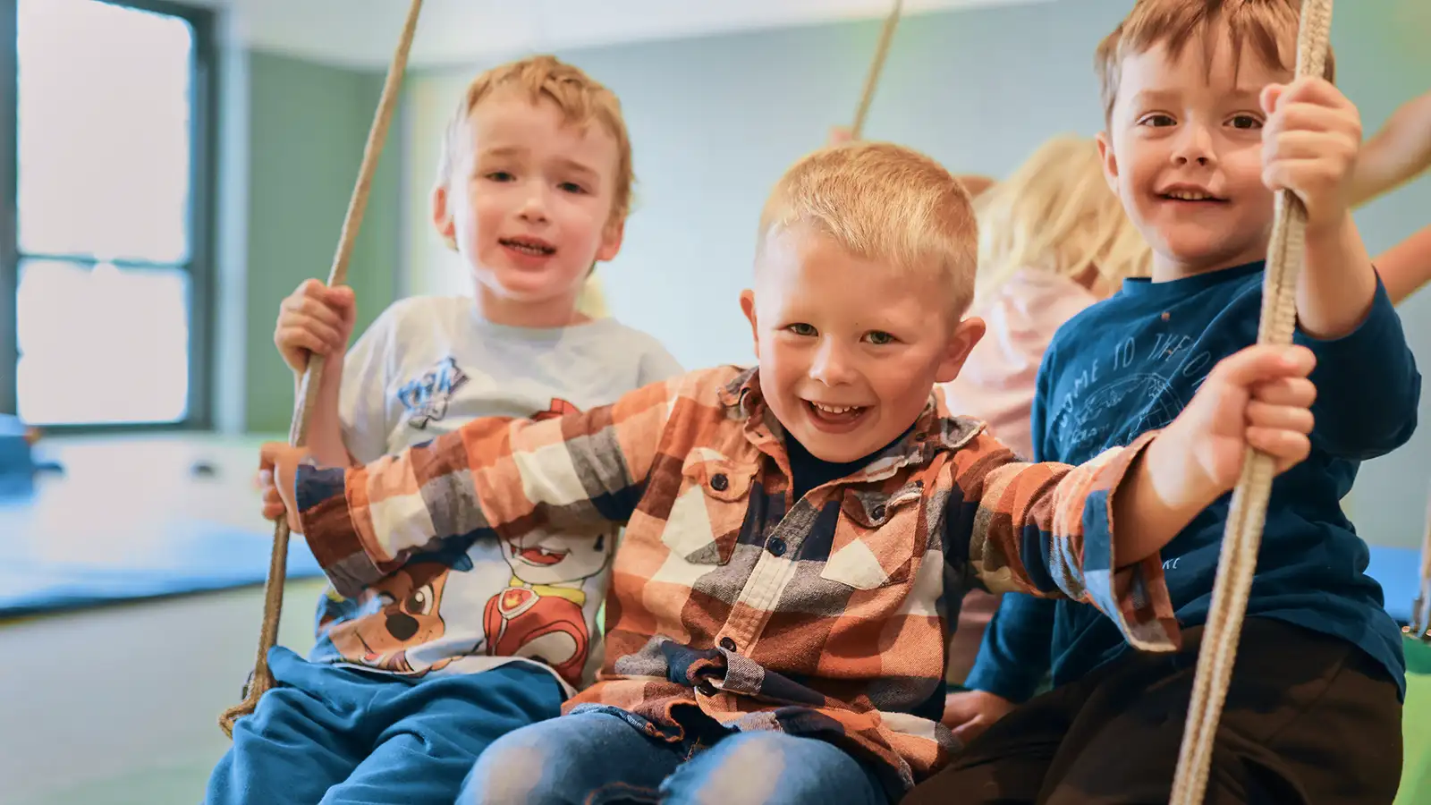 Drei lachende Kinder schaukeln zusammen auf einer Seilschaukel in einem Spielzimmer. Sie tragen bunte Kleidung und haben sichtlich Spaß.