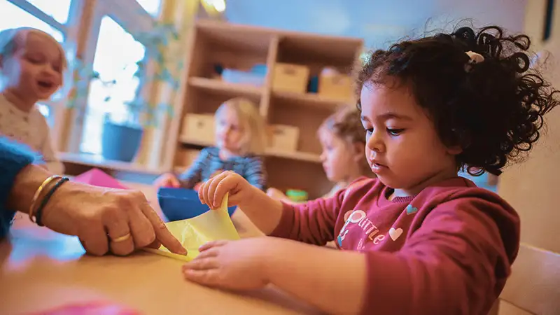 Ein Kind bastelt konzentriert im Kindergarten, während drei weitere Kinder im Hintergrund spielen. Ein Erwachsener hilft.