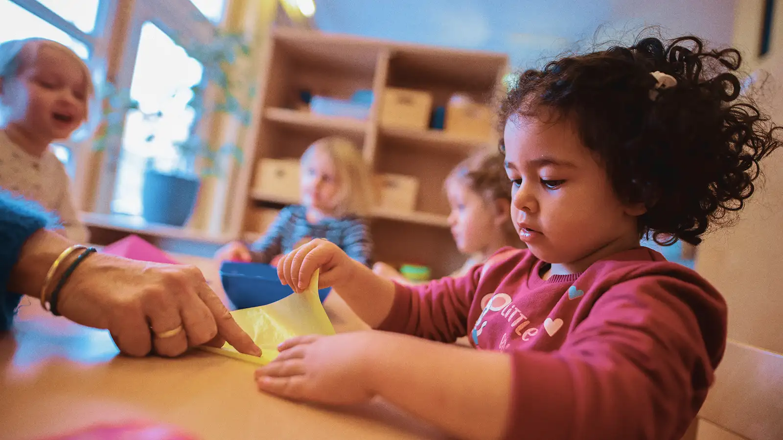 Ein Kind bastelt konzentriert im Kindergarten, während drei weitere Kinder im Hintergrund spielen. Ein Erwachsener hilft.