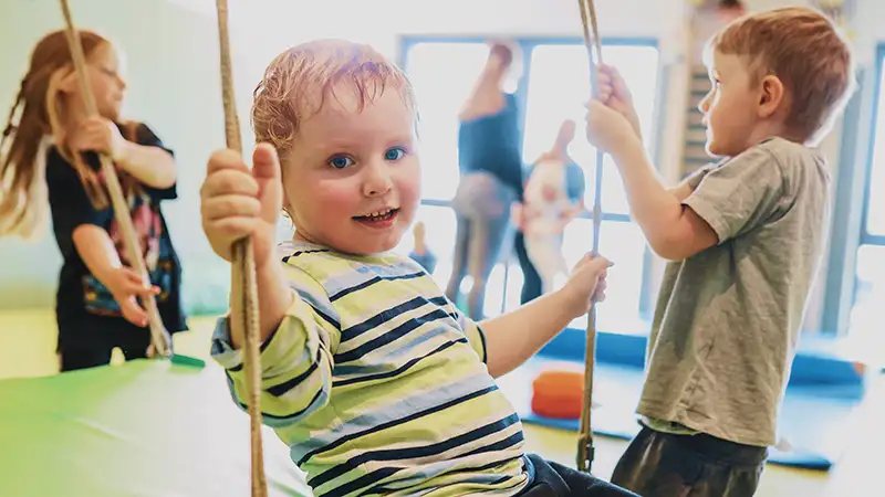 Ein Kind schaukelt im Kindergarten, während ein anderes Kind daneben steht. Im Hintergrund sind weitere Kinder zu sehen.