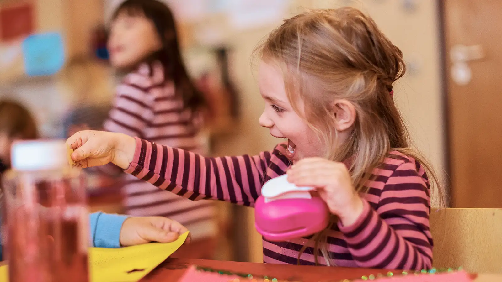 Ein lachendes Mädchen in einem Kindergarten bastelt mit Papier und Klebstoff, während andere Kinder im Hintergrund spielen.