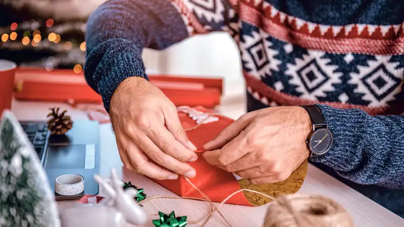 Nahaufnahme von Händen, die ein Geschenk in rotes Geschenkpapier wickeln. Die Person trägt einen weihnachtlichen Pullover.