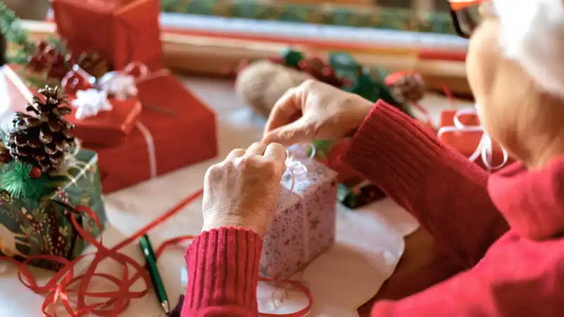 Nahaufnahme von Händen einer Person, die ein Geschenk mit einer Schleife verziert. Im Hintergrund sind weitere Geschenke und Dekorationen zu sehen.