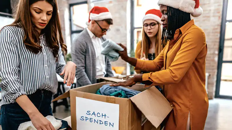 Vier Personen mit Weihnachtsmützen stehen um einen Karton mit dem Aufdruck „Sachspenden“ und legen Kleidung hinein.