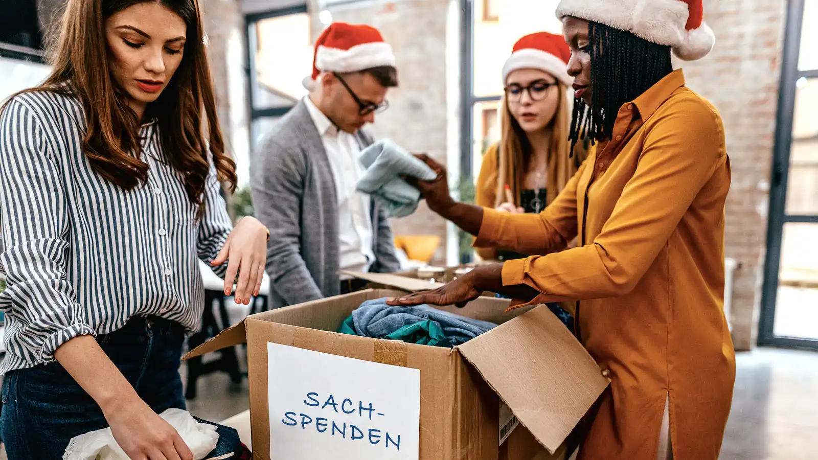 Vier Personen mit Weihnachtsmützen stehen um einen Karton mit dem Aufdruck „Sachspenden“ und legen Kleidung hinein.