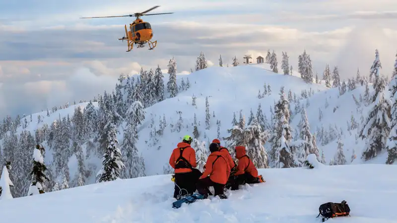 Rettungsteam in Schneeausrüstung sitzt auf einem Berggipfel, ein Helikopter schwebt über ihnen.