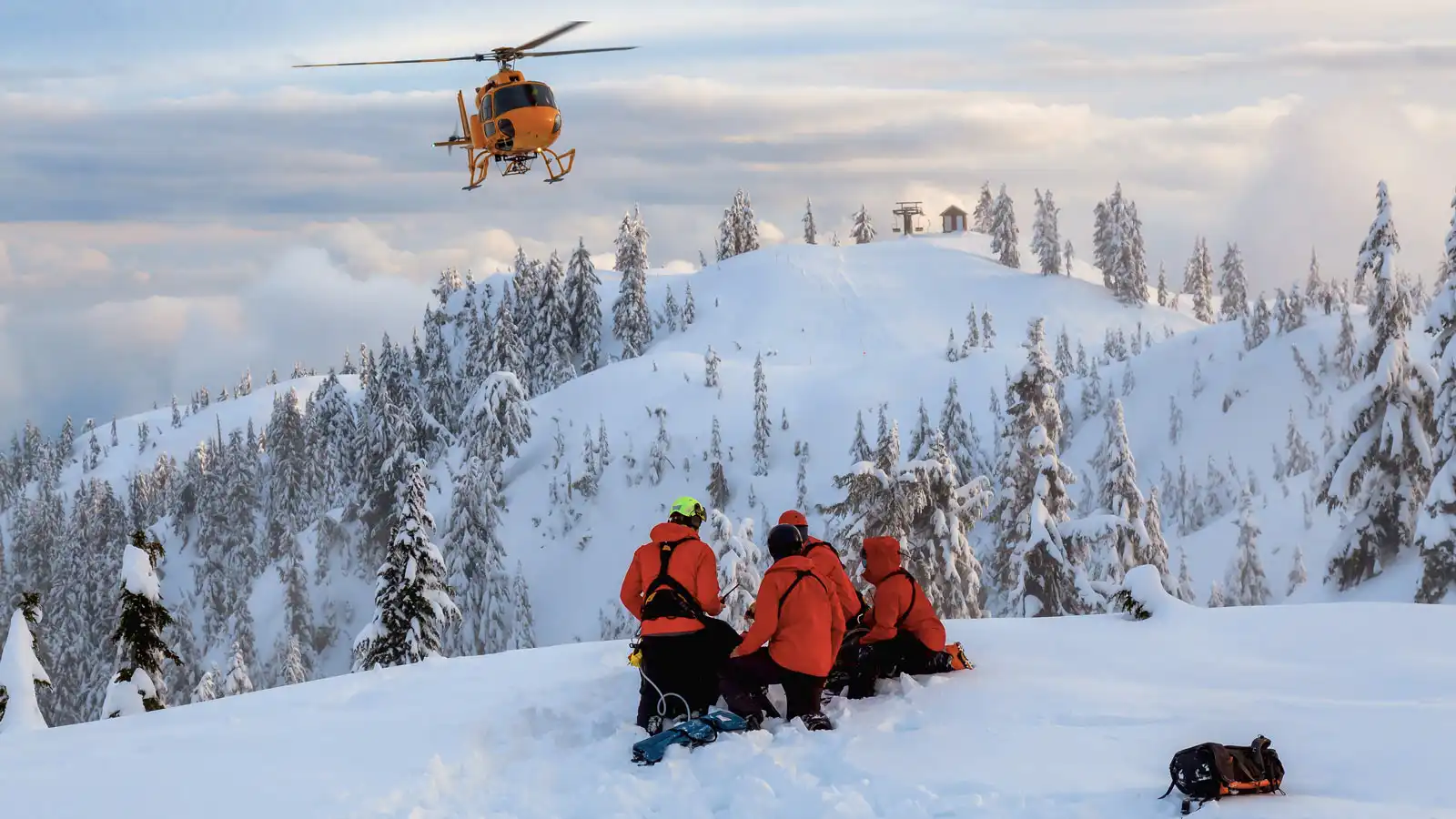 Rettungsteam in Schneeausrüstung sitzt auf einem Berggipfel, ein Helikopter schwebt über ihnen.