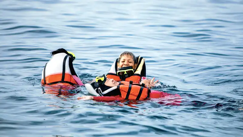 Drei Menschen in Rettungswesten schwimmen im Wasser, einer stützt einen anderen.