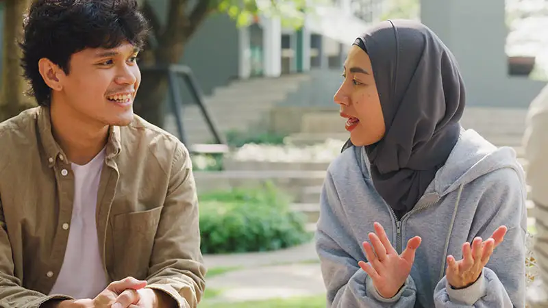 Zwei Personen sitzen im Freien und unterhalten sich. Eine trägt einen Hijab und gestikuliert mit den Händen, während die andere lächelt und zuhört.