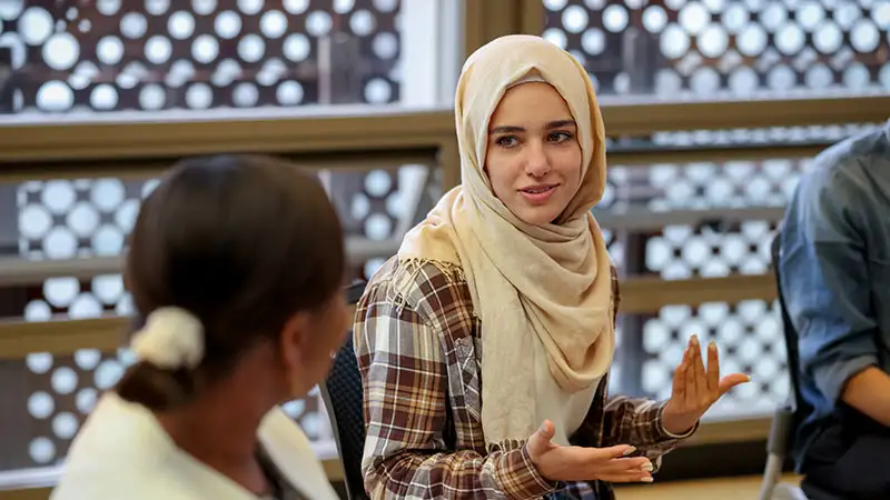 Eine Person trägt einen hellen Hijab und gestikuliert während eines Gesprächs mit einer anderen Person.