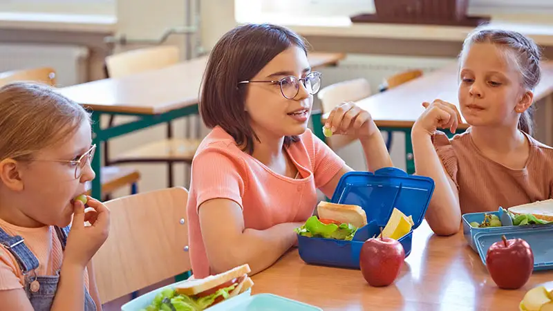 Drei Schülerinnen sitzen an einem Tisch und essen aus ihren Schultüten. Sie essen Obst, Salat und Sandwiches.