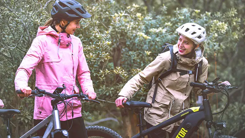 Zwei Radfahrerinnen im Freien, beide mit Helmen und Fahrrädern, lächelnd und in einer entspannten Pose.
