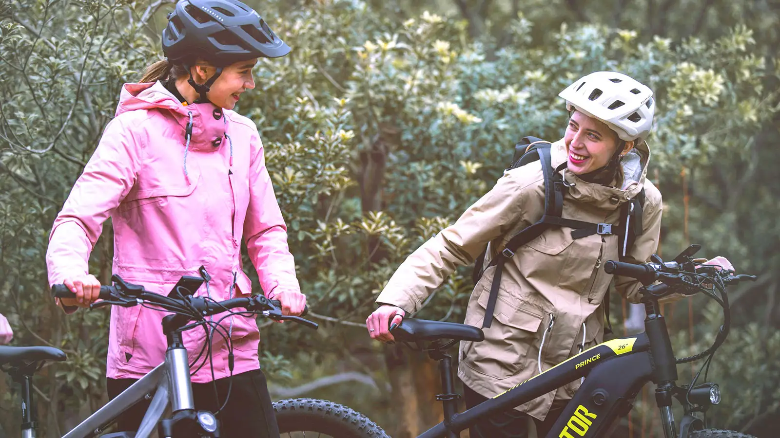 Zwei Radfahrerinnen im Freien, beide mit Helmen und Fahrrädern, lächelnd und in einer entspannten Pose.