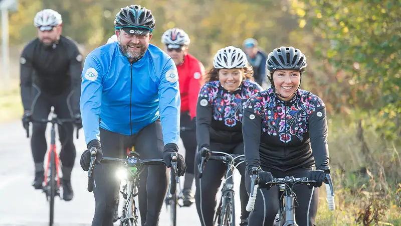 Gruppe von Radfahrern auf einer Straße, alle mit Helmen und sportlicher Kleidung, einer davon lächelt in die Kamera.