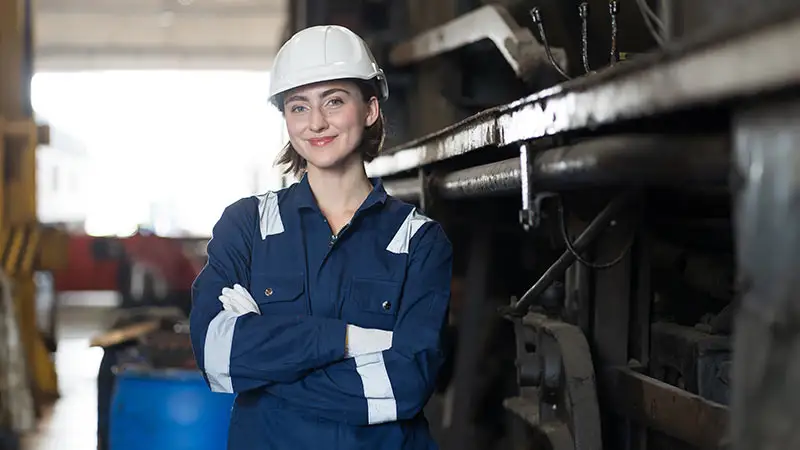 Eine Person in blauer Arbeitskleidung und weißem Helm steht in einer Industriehalle mit verschränkten Armen.