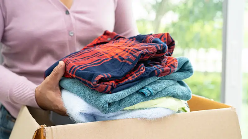 Eine Person hält eine gestapelte Kleidung aus blauen und orangefarbenen Stoffen in einer braunen Kartonbox.