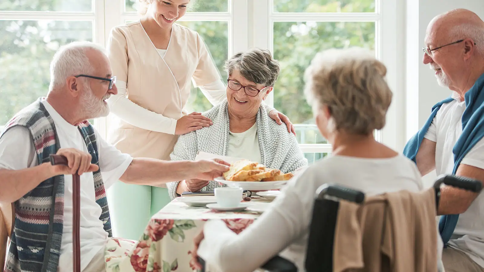 Eine Pflegekraft hilft älteren Menschen beim gemeinsamen Frühstück an einem Tisch.